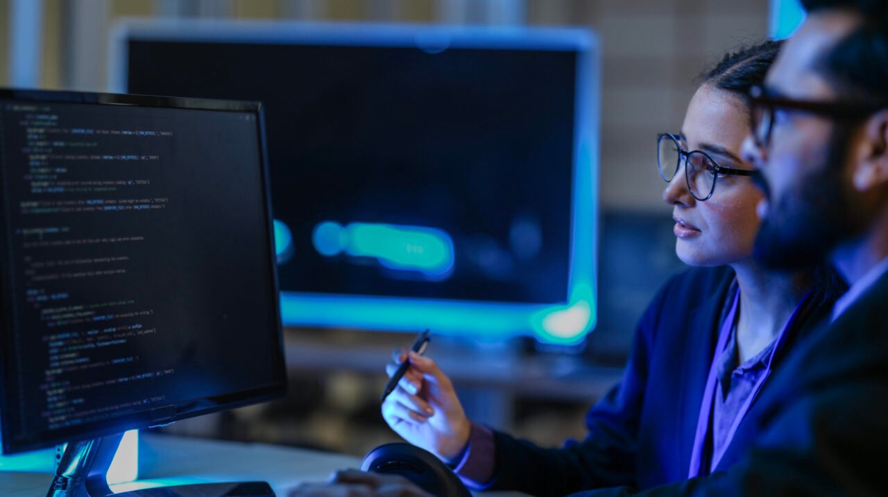 diverse AI developers collaborate in a late-night coding session. A senior engineer mentors his colleague as they program a complex machine learning algorithm together.
